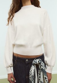 Maglione crema tagliato con collo alto e maniche a sbuffo, abbinato a una gonna di jeans scura con una sciarpa decorata legata in vita.