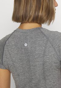 Siva atletska majica s teksturiranim uzorkom, okruglim izrezom i kratkim raglan rukavima. Ima mali logo na leđima.