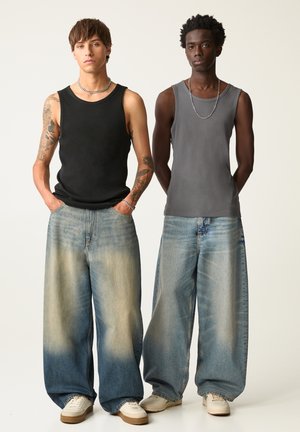 Deux modèles se tiennent côte à côte, portant des débardeurs ; l'un est en noir, l'autre en gris. Tous deux portent des jeans délavés à jambes larges et des baskets.