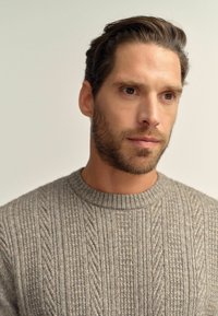 Gray cable-knit sweater featuring ribbed neckline, shown on a model with dark hair and a light background, showcasing texture and pattern details.