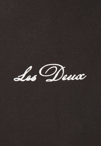 Black fabric with a textured surface featuring the embroidered text "Les Deux" in white cursive font, positioned centrally.