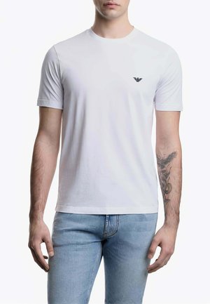 Uomo che indossa una maglietta bianca semplice a maniche corte con un piccolo logo nero sul petto e jeans azzurri, con un tatuaggio sull'avambraccio destro.