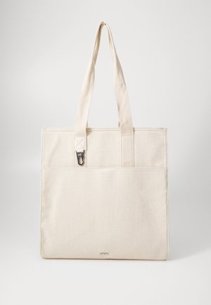 Sac cabas en toile beige avec deux longues anses, poche extérieure avant et petite pince métallique fixée près de la anse.