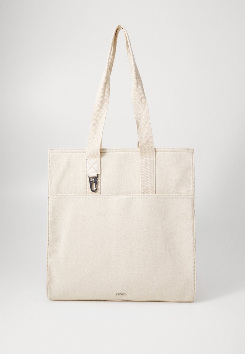 Sac cabas en toile beige avec deux longues anses, poche extérieure avant et petite pince métallique fixée près de la anse.