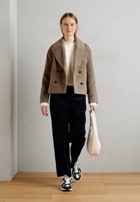 Vrouw die binnen loopt in een bruine double-breasted jas, witte trui, zwarte broek, zwart-witte sneakers en een witte tas draagt.