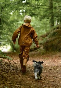 Kind in een bruine waterdichte jas en bijpassende broek loopt over een bospaadje met een kleine, grijze hond. Weelderig groen gebladerte omringt hen.