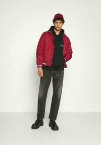 Bordeaux bomberjakke over en sort hættetrøje, parret med grå jeans og sorte sko. Jakken har ribbede detaljer og lynlås lukning.