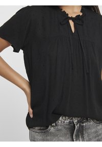 Blouse noire à manches courtes avec un col à volants et un détail noué. Le tissu texturé présente de petits points en relief et une coupe décontractée.
