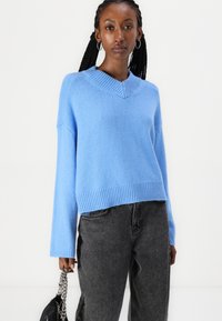 Lyseblå strikkegenser med V-hals, ribbestrikket nederkant og lange ermer. Kombinert med grå jeans og holder en svart veske med metallkjede.