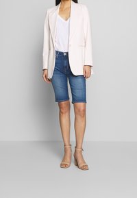 Un blazer rosa chiaro, una maglietta bianca a scollo a V, pantaloni bermuda in denim blu e sandali beige con cinturini presentano un look informale ma curato.