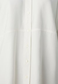 Camicia bianca con superficie texturizzata, caratterizzata da un taglio dritto e una placca a mezza bottoni. Design semplice senza motivi.