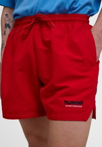 Röda badshorts i lätt material, med elastisk midja och snörning samt sidofickor. Svart logotyp på nederkanten.