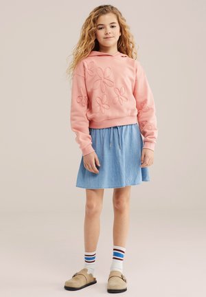 Ragazza giovane con capelli ricci che indossa una felpa con cappuccio ricamata con fiori rosa, gonna blu, calze a righe e scarpe slip-on beige.