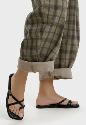 Voeten met zwarte platte sandalen met bandjes, gecombineerd met opgerolde beige en groene geruite broek tegen een effen achtergrond.