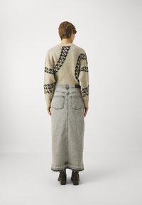 Pull beige avec motif de chaînes noires, associé à une longue jupe en denim grise et des bottines noires. Fond uni, vue de dos.