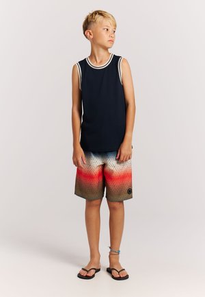 Navy mouwloos topje met witte bies, gecombineerd met multicolor zwemshorts met een geometrisch patroon in rood, oranje en beige. Zwarte teenslippers.