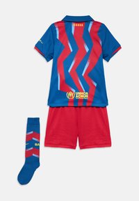 Echipament de fotbal pentru copii, cu un tricou albastru și roșu, decorat cu modele zigzag și logo-uri, asortat cu pantaloni scurți roșii și șosete albastre și roșii.