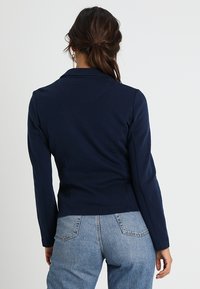 Marinblå kavaj med slagkrage, skräddarsydd sömmar och långa ärmar, i kombination med ljusblå högmidjade jeans.