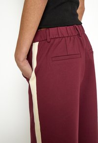 Burgundy pants with an elastic waistband, featuring cream side stripes and back pockets. Smooth fabric with a casual fit.