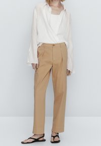 Chemise blanche en lin légère à manches longues, associée à un pantalon plissé taille haute couleur beige et des sandales plates noires. Design et coupe minimalistes.