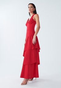 Red sleeveless maxi dress with a v-neckline, layered ruffles at the skirt, and a smooth, slightly sheer fabric. Simple strappy heels.