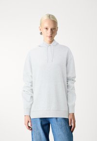 Lichtgrijze hoodie van zacht materiaal met een kangoeroezak. Voorzien van een capuchon met trekkoord en lange, brede mouwen. Gecombineerd met blauwe spijkerbroek.