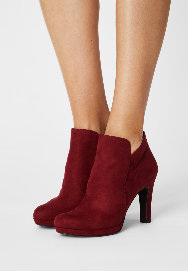 Bas des jambes d'une femme portant des bottines en daim rouge à talons hauts avec bouts arrondis sur un fond neutre.
