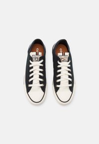 Czarne trampki na płaskiej podeszwie z białą gumową podeszwą i czubkiem. Posiadają białe sznurówki i logo "Converse All Star" na języku. Dyskretne detale tekstury.