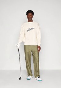 Krémová mikina s nápisom "Malbon" v modrom písme, v kombinácii s olivovozelenými nohavicami a bielymi golfovými topánkami. Postava drží golfovú palicu.