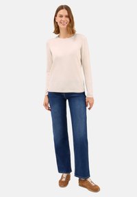 Giovane donna in piedi che indossa un maglione beige chiaro a maniche lunghe, jeans blu a gamba dritta e sneakers marroni su uno sfondo neutro.