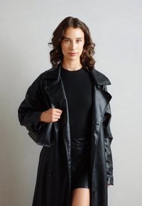 Must negra kunstnahk jooksjakott laia krae, kahekordse nööbiga ja läikiva tekstuuriga, kombineeritud musta soonikkoes toppi ja lühikeste pükstega.