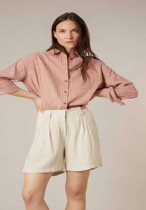 Femme aux longs cheveux bruns portant une chemise ample à rayures roses et blanches boutonnée et un short beige taille haute, debout les mains sur les hanches.