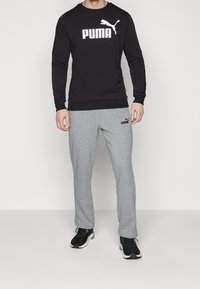 Hombre que lleva puesto un sudadera negra de Puma y pantalones deportivos grises de Puma con zapatillas deportivas en blanco y negro, de pie contra un fondo liso.
