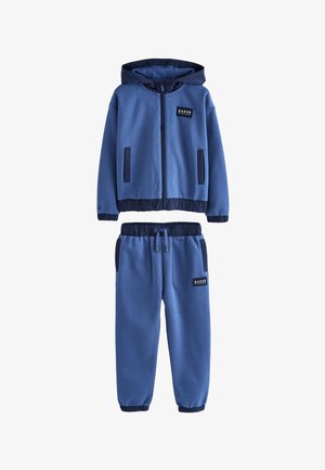 Kék két részes tréningruha kapucnis cipzáras dzsekivel és rugalmas derékpántos, húzózsinórral ellátott nadrággal, fekete díszítéssel és "Baker by Ted Baker" logóval ellátott foltokkal.