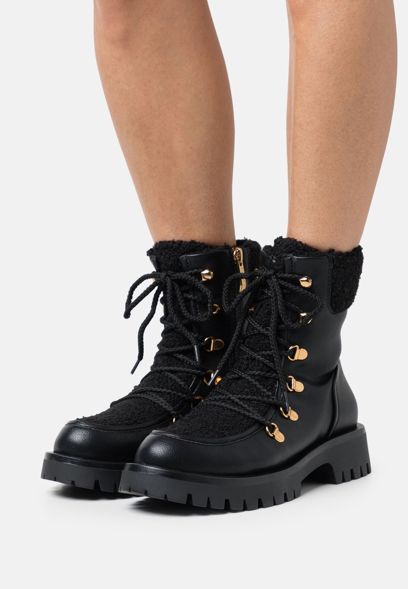 Bottes noires avec doublure en fausse fourrure, tissu texturé, quincaillerie dorée et semelles épaisses dotées de profonds crampons. Design à lacets avec fermeture éclair sur le côté.