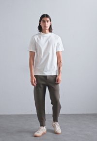 Tânăr stând, îmbrăcat cu o cămașă albă cu mâneci scurte, pantaloni jogger închiși la culoare și adidași albi, având brațele tatuate, pe un fundal gri simplu.