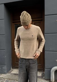 Muž v béžovom pletenom svetri a zodpovedajúcej čiapke sa pozerá dole s rukami pri vreckách, stojaci pred drevenými dverami a sivou stenou.