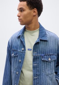 Νέος άνδρας με μπλε ριγέ τζιν μπουφάν πάνω από ένα ανοιχτό πράσινο πουκάμισο με λογότυπα "HUGO", κοιτάζει προς τα αριστερά πάνω σε απλό φόντο.