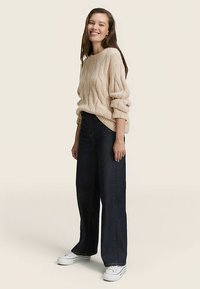 Pull en maille beige avec des manches surdimensionnées, associé à un jean large bleu foncé et des baskets blanches, sur un fond neutre.