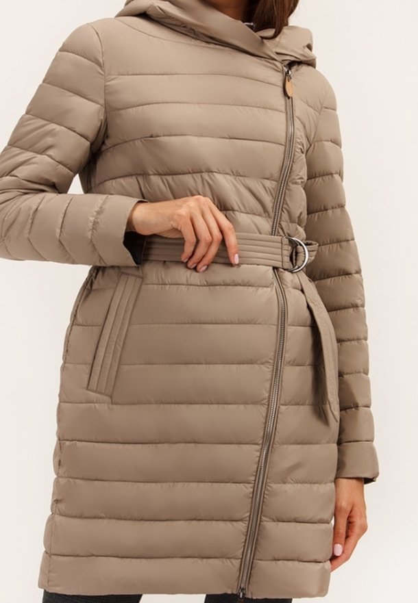 Žena oblečená v béžovém prošívaném péřovém kabátu s kapucí a páskem v pase, upravuje pásek levou rukou.