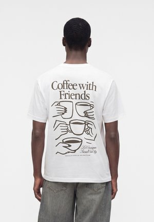 Mann trägt weißes T-Shirt mit dem Text "Coffee with Friends" und Illustrationen von Händen, die Kaffeetassen auf dem Rücken halten.