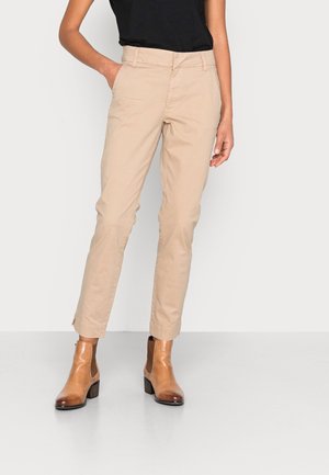 Frau, die beige gerade geschnittene Hose, schwarzen Top und hellbraune Lederknöchelstiefel trägt, steht mit einer Hand in der Tasche.