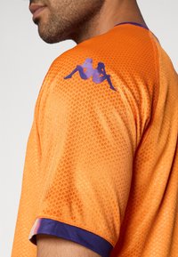 Tricou sport portocaliu cu un model texturat în formă de grilaj, având margini violet pe guler și un logo grafic violet pe umăr.