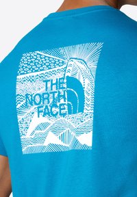 Camiseta de algodón turquesa con un gran gráfico blanco que presenta patrones de líneas abstractas y el texto "THE NORTH FACE" centrado en la parte posterior.
