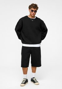 Zwart oversized sweatshirt, witte t-shirt eronder, zwarte shorts en zwarte sneakers met crèmekleurige accenten. Krullend haar, draagt donkere zonnebril.