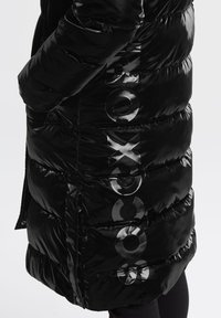 Schwarze Pufferjacke mit glänzendem, gestepptem Design, auffälliger Markenbeschriftung in glänzendem Text an der Seite und entspannter Silhouette.