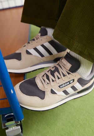 Beigen ja mustan väriset Adidas-tennarit, joissa valkoiset raidat, valkoisten sukkien ja vihreiden samettihousujen kanssa, lepäävät vihreällä lattialla lähellä sinistä metallikehystä.