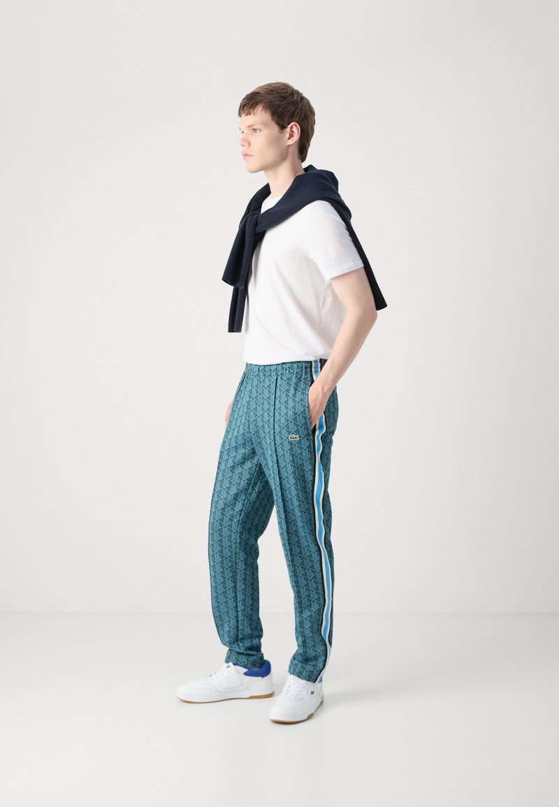 Pantalon de jogging teal à motifs avec des rayures latérales blanches et bleues et un logo brodé ; associé à un t-shirt blanc et un pull marine drapé sur les épaules.