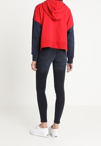 Mujer con una sudadera roja y azul marino, jeans ajustados oscuros y zapatillas blancas, de pie con la espalda hacia la cámara sobre un fondo liso.