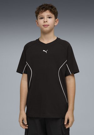 Niño con cabello rizado y corto, vistiendo una camiseta negra de Puma con ribetes blancos, de pie contra un fondo gris liso.